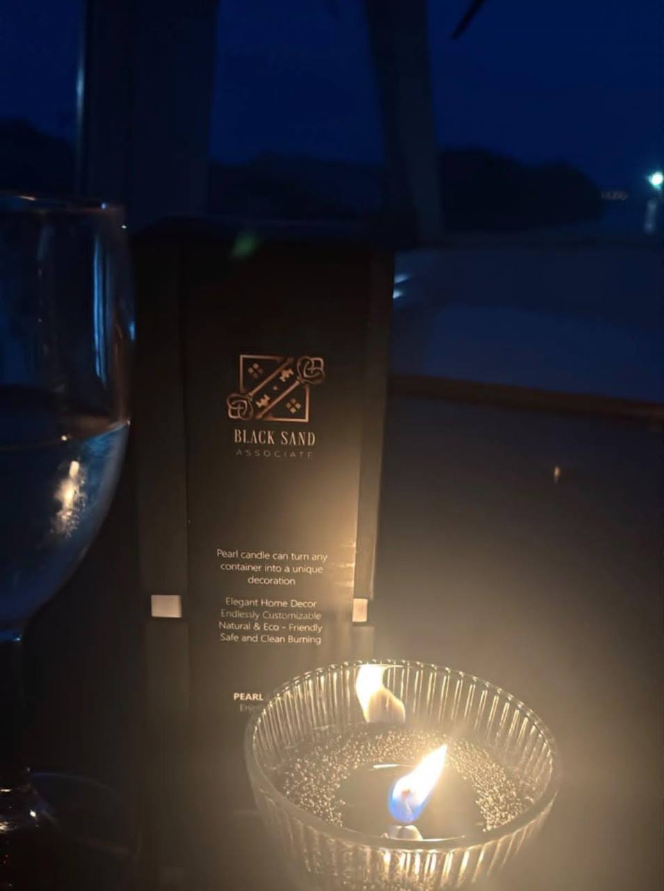 Black Sand pearl candle glowing in a fluted glass vessel at dusk